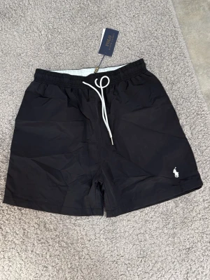 Svarta badbyxor från Polo Ralph Lauren - Snygga svarta badbyxor från Polo Ralph Lauren med vit dragsko i midjan och klassisk logga broderad på benet. Tillverkade i snabbtorkande material och har meshfoder på insidan för extra komfort. Perfekta för stranden eller poolen.