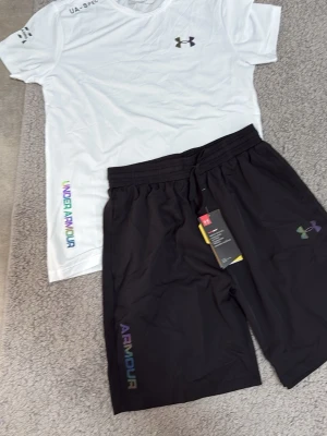 Svarta shorts från Under Armour - Säljer ett par svarta shorts från Under Armour med regnbågsskimrande logga och text på benet. Shortsen har snörning i midjan och är tillverkade i ett lätt och svalt syntetmaterial, perfekta för träning eller chill dagar.