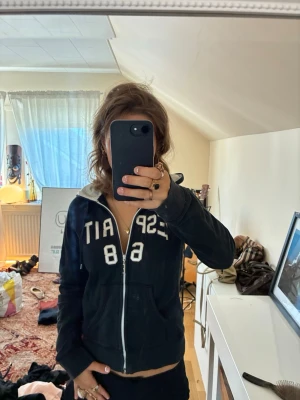Mörkblå hoodie med dragkedja från esprit - Säljer en mörkblå hoodie med vit dragkedja och ett stort vitt esprit-märke på bröstet. Hoodien har huva med snören och två fickor framtill. Perfekt för en avslappnad och sportig stil.
