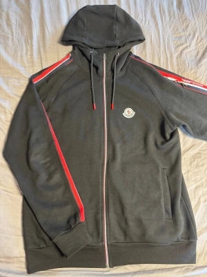 Svart Moncler hoodie med röd detalj - Snygg svart hoodie från Moncler med dragkedja, huva och röda samt vita ränder längs ärmarna. Klassisk Moncler-logga på bröstet och röda detaljer på snörena. Tillverkad i mjukt bomullsmaterial och har två fickor framtill.