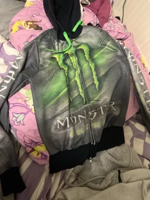 Monster Energy zip hoodie grå/svart - Säljer en grå och svart zip hoodie med stort grönt Monster Energy-tryck på bröstet och text på ärmarna. Huvan har gröna snören och insidan är svart. Perfekt för dig som gillar streetwear och vill sticka ut med en cool design.