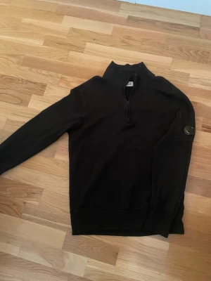 Svart half-zip tröja från C.P. Company - Svart långärmad tröja från C.P. Company med half-zip och hög krage. Tröjan har den ikoniska linsen på ärmen och är tillverkad i mjukt material, perfekt för en avslappnad och stilren look.