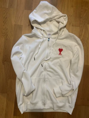 Vit hoodie med röd Ami-dekal - Säljer en vit hoodie från Ami med dragkedja, huva och snörning. På bröstet finns Ami:s ikoniska röda hjärta med bokstaven A. Hoodien har fickor framtill och ribbade muddar vid ärmar och nederkant. Perfekt för en clean och stilren look.
