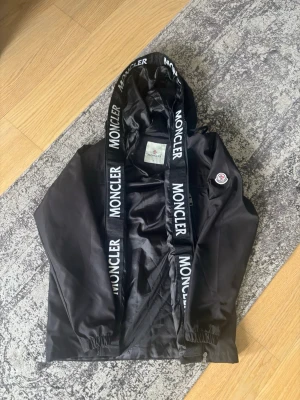 Svart vindjacka med huva - Snygg svart vindjacka från med stor huva och breda logoband. Jackan har två dragkedjeförsedda fickor framtill, elastiska muddar vid ärmslut och logga på ärmen. Materialet är tunt och vindtätt, perfekt för blåsiga dagar. Priset kan diskuteras!!