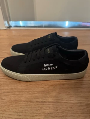 Svarta Saint Laurent sneakers - Snygga svarta sneakers från Saint Laurent med vit text på sidan och gulddetaljer på plösen. Skorna har en klassisk låg profil, svarta skosnören och vit sula. Materialet ser ut att vara en mix av mocka och textil för en stilren look.