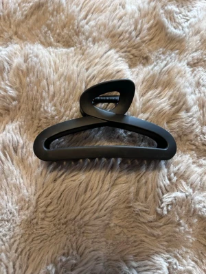 Svart hårklämma i matt plast - Stilren svart hårklämma i matt plast med triangulär form och bred öppning. Klämman har stark fjäder och greppvänliga tänder som håller håret på plats hela dagen. Perfekt accessoar för att snabbt fixa frisyren.