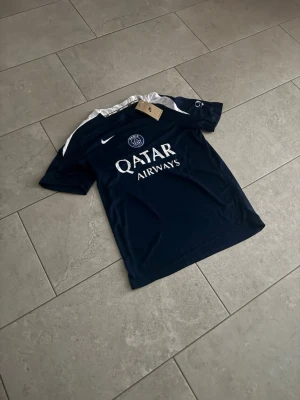 Mörkblå PSG matchtröja från Nike - Snygg mörkblå PSG matchtröja från Nike med vita detaljer på axlarna och klubbmärke på bröstet. Framsidan har Qatar Airways-tryck och tröjan är tillverkad i ett lätt och funktionellt material som passar perfekt till fotbollsträning eller match.