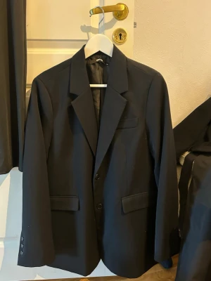 Svart kavaj med klassisk passform - Stilren svart kavaj med två knappar framtill, klassisk krage och tre fickor. Kavajen har slät yta och är helfodrad, vilket ger en snygg och tidlös look. Perfekt för dig som vill ha en enkel men elegant kavaj till garderoben.