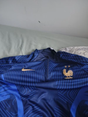Blå Nike Frankrike träningströja - Snygg blå långärmad träningströja från Nike med Frankrikes landslagsmärke och två stjärnor på bröstet. Tröjan har halv dragkedja framtill och tunna blå linjemönster över hela plagget. Perfekt för fotbollsträning eller sport.