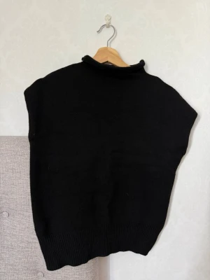 Svart stickad väst med hög krage - En svart stickad väst med hög krage och ribbad nederkant. Västen har en enkel och stilren design utan ärmar, perfekt att bära över en skjorta eller tröja för en trendig lager-på-lager-look.