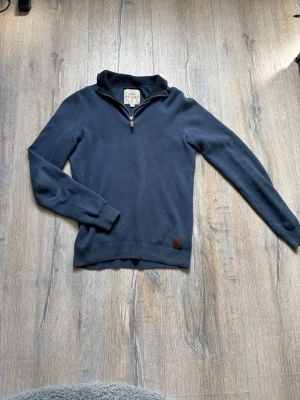 Mörkblå halvzip tröja Next Supply - Mörkblå stickad tröja från Next Supply med halvzip och ståkrage. Tröjan har långa ärmar, ribbade muddar och en liten läderdetalj vid nederkanten. Perfekt för lager på lager och en clean look.