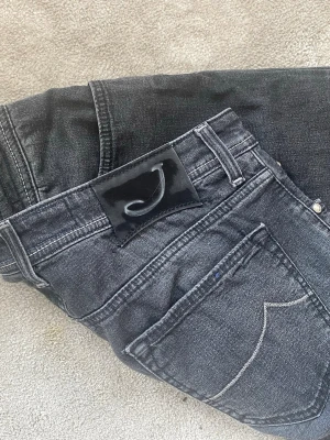 Svarta Jacob Cohen Jeans - Säljer ett par svarta tvättade jeans med raka ben och kontrastsömmar på bakfickorna. Jeansen har en svart patch med ett J på linningen och klassisk femficksdesign. Perfekta för en avslappnad och trendig look. 