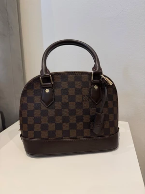 Brun handväska - Säljer en klassisk handväska i brunt med två handtag och guldfärgade metalldetaljer. Väskan har en rundad form och detaljer i mörkbrunt läder. Perfekt för dig som gillar snygga handväskor och tidlös design. Helt ny.