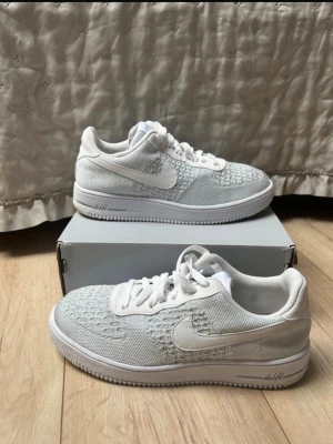 Vita Nike Air Force 1 sneakers - Säljer ett par fräscha vita Nike Air Force 1 sneakers med stickad ovandel och klassisk snörning. Skorna har en tydlig Nike swoosh på sidan och en bekväm sula med AIR-detalj. Perfekta för dig som gillar stilrena och sportiga sneakers.