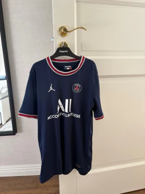 Mörkblå PSG matchtröja 2020 - 2021 Jordan - Säljer en mörkblå PSG matchtröja med vita och röda detaljer vid krage och ärmslut. Tröjan har PSG-logga och Jordan-logga på bröstet samt sponsortryck framtill. Tillverkad i lätt och ventilerande material, perfekt för fotboll eller träning.