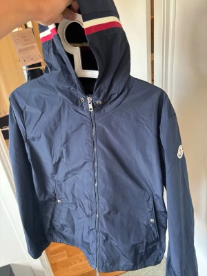 Marinblå vindjacka från Moncler - Snygg marinblå vindjacka från Moncler med huva och dragkedja framtill. Jackan har vita och röda ränder på huvan samt Moncler-logga på ärmen. Två fickor med tryckknappar och lätt, vindtätt material. Perfekt för blåsiga dagar.