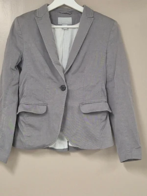 Grå kavaj från H&M, storlek 36 - Snygg grå kavaj från H&M i storlek 36. Klassisk modell med en knapp framtill, två fickor med lock och smal krage. Fodrad med randigt tyg på insidan.