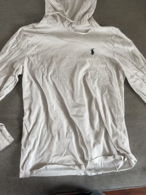 Vit hoodie från Polo Ralph Lauren - Säljer en vit hoodie från Polo Ralph Lauren med den klassiska loggan broderad på bröstet. Hoodien har huva med dragsko och är tillverkad i ett mjukt och tunt bomullsmaterial. Perfekt för en clean och avslappnad stil.