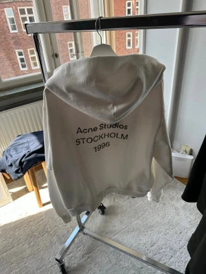 Vit hoodie från Acne Studios - Säljer en vit hoodie från Acne Studios med tryck på ryggen där det står 'Acne Studios STOCKHOLM 1996'. Hoodien har en klassisk passform, huva och ribbade muddar. Perfekt för en clean och stilren look. Äkta ! Modellen är 187cm
