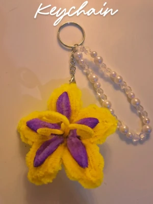 Tangled Keychain - Unik nyckelring med en handgjord blomma i gult och lila av piprensare. Nyckelringen har en kedja och ett band av vita och genomskinliga pärlor. Perfekt som accessoar till väskan eller nycklarna.