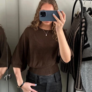 Brun stickad tröja med ballongärm - Mysig brun stickad tröja med bred ribbad midja och ballongärmar i trekvartsmodell. Tröjan har rund halsringning och är tillverkad i ett mjukt, tunt material som ger en avslappnad men ändå stilren look.