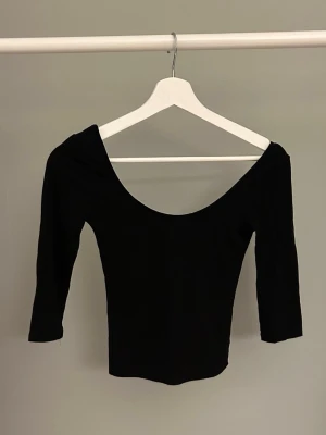 Svart offshoulder topp med trekvartsärm - Snygg svart offshoulder topp med djup urringning både fram och bak. Toppen har trekvartsärm och en tight passform som framhäver figuren. Perfekt att styla med höga jeans eller kjol för en trendig look.