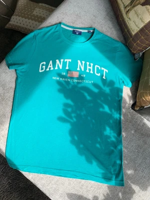 Turkos GANT t-shirt med tryck - Snygg turkos t-shirt från GANT med vit text och amerikansk flagga på bröstet. Klassisk rund hals och korta ärmar. Perfekt för dig som gillar college-vibe och vill ha en enkel men stilren look.