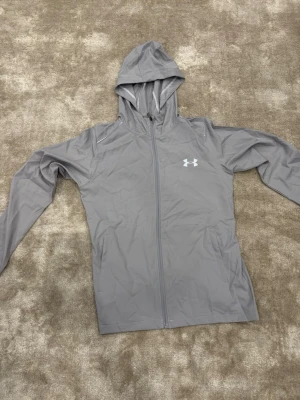 Under Armour HeatGear jacka storlek M/S -  Snygg och lätt Under Armour-jacka i grått med huva och reflexdetaljer som syns tydligt i mörker. Perfekt för träning, löpning eller vardagsbruk. HeatGear-material som känns svalt, torrt och lätt. Stilren och sportig! 🔥  Storlek: M passar även i S Märke: Under Armour