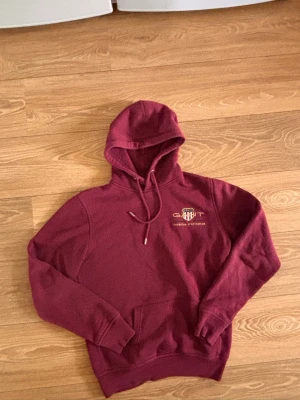 Vinröd hoodie från GANT  - Säljer en vinröd hoodie från GANT med huva och dragsnören. På bröstet finns ett broderat GANT-logotyp och texten 'American Sportswear'. Hoodien har magficka och ribbade muddar. Perfekt för dig som gillar klassisk streetwear.