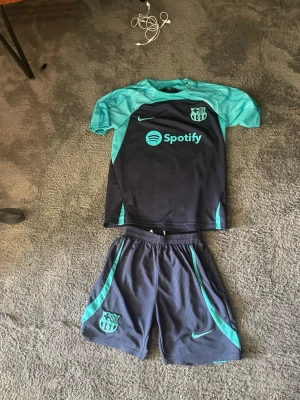 Barcelona träningsset Nike blå/turkos - Säljer ett FC Barcelona träningsset från Nike med kortärmad tröja och shorts. Setet är mörkblått med turkosa detaljer och loggor, samt Spotify som sponsortryck. Perfekt för fotbollsträning eller gymmet.