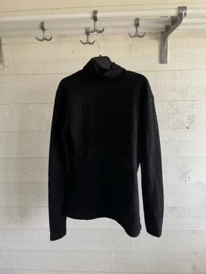Svart polotröja från Amisu - Säljer en svart polotröja från Amisu med diskret struktur i tyget. Tröjan har lång ärm och hög krage, perfekt för lager på lager. Enkel och stilren design som passar till det mesta.
