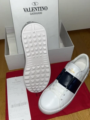 Valentino Garavani vita sneakers - Snygga vita sneakers från Valentino Garavani med bred mörkblå rem över vristen och guldfärgad logga på plösen. Skorna har en chunky sula med nitar och är tillverkade i slätt läder. Perfekta för dig som vill ha en exklusiv och trendig look.