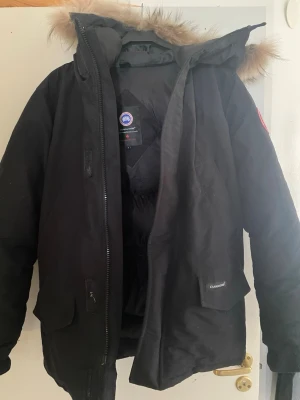 Svart dunjacka med pälskant - Säljer en svart dunjacka från Canada Goose med huva och avtagbar pälskant. Jackan har dragkedja, stora fickor och klassisk patch på ärmen. Perfekt för kalla vinterdagar och riktigt varm tack vare dunfyllningen.