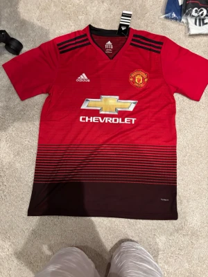 Manchester United 18/19 #pogba 6 - Röd och svart Manchester United matchtröja från Adidas med Pogba och nummer 6 på ryggen. Tröjan har klubbmärke, Adidas-logga och Chevrolet som sponsor framtill. Tillverkad i lätt och ventilerande material, perfekt för fotbollsträning eller match.