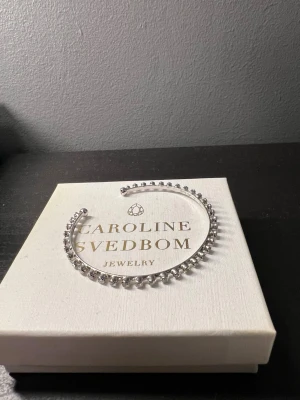 Silverfärgat armband Caroline Svedbom - Snyggt armband i silverfärg från Caroline Svedbom. Armbandet är stelt och dekorerat med gnistrande, klara pärlor i rad längs hela bandet. Perfekt för dig som gillar stilrena och glittriga accessoarer. Aldrig använd 