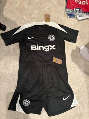 Chelsea 26/27 träningsställ - Säljer ett svart Chelsea träningsset från Nike med vita detaljer. Tröjan har BingX-logga och klubbmärke på bröstet samt vit Nike-logga. Shortsen har elastisk midja, klubbmärke och vit Nike-logga. Perfekt för fotbollsträning eller gymmet.