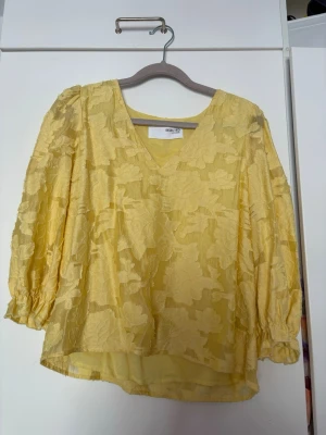 Gul blus med blommönster och v-ringning - Snygg gul blus med v-ringning och trekvartsärm. Blusen har ett upphöjt blommönster i tyget som ger en fin struktur och en luftig känsla. Perfekt att styla med jeans eller kjol för en trendig look.