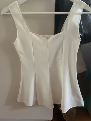Vit figurnära linnetopp - Säljer en stilren vit linnetopp med breda axelband och lätt utsvängd nederkant. Toppen har en figurnära passform och en subtil v-ringning framtill. Perfekt att matcha med jeans eller kjol för en fräsch look.