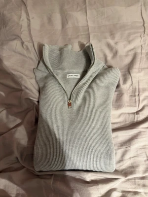 Ljusgrå ribbstickad tröja Jack & Jones - Ljusgrå ribbstickad tröja från Jack & Jones med halv dragkedja och hög krage. Tröjan har en clean look och är tillverkad i mjukt material som känns skönt mot huden. Perfekt för lager på lager och enkel att matcha med olika outfits.