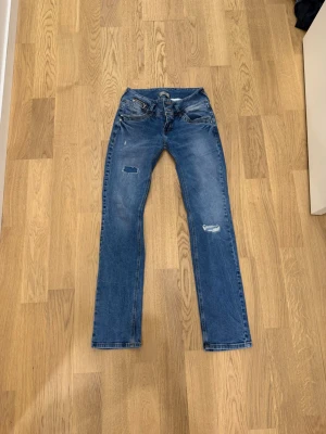 Blå raka jeans med slitningar - Säljer ett par blå jeans som är från LTB med raka ben och coola slitningar på låret och knät. Jeansen har dubbla knappar i midjan, fem fickor och dragkedja på myntfickan. Perfekta för en avslappnad och trendig look. Priset kan diskuteras vid snabb affär 