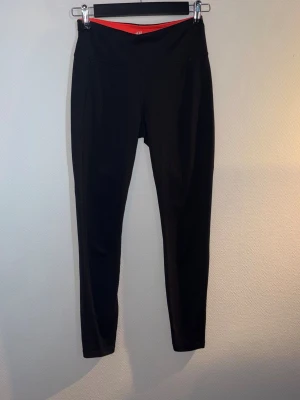 Svarta leggings med hög midja - Säljer ett par svarta leggings med hög midja och smal passform. De har en diskret detalj med orange insida i linningen. Perfekta till både träning och chill. Materialet känns stretchigt och mjukt. Storlek S men sitter som XS.