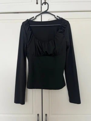 Svart långärmad topp med rynkad urringning - Stilren svart topp med långa ärmar och en snygg rynkad urringning. Modellen är figurnära och har en klassisk, enkel design som passar till många olika outfits.