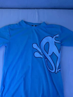 Blå t-shirt med grafiskt tryck syna world - Snygg blå t-shirt med stort grafiskt tryck i svart och vitt både fram och bak. Klassisk rund hals och korta ärmar. Perfekt för dig som gillar streetwear och vill sticka ut lite extra.