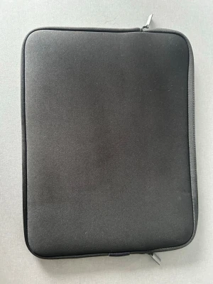 Svart laptopfodral med dragkedja - En stilren svart datorväska i mjukt material med dragkedja runt kanten. Fodralet har en enkel, rektangulär form och passar perfekt för att skydda din laptop eller surfplatta. Praktisk och smidig att bära med sig till skolan eller jobbet.