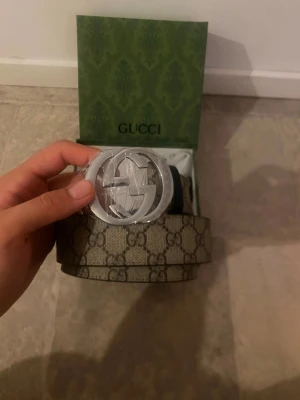 Gucci beige bälte med GG-spänne - Säljer ett snyggt beige bälte från Gucci med det klassiska GG-mönstret och ett stort, silverfärgat GG-spänne. Bältet kommer i originalförpackning och har en exklusiv känsla som lyfter vilken outfit som helst.