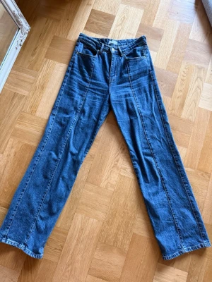 Blå raka jeans med pressveck - Säljer ett par blå jeans med raka ben och markerade pressveck framtill. Klassisk femficksmodell med hög midja och bälteshällor. Jeansen har en avslappnad passform och är tillverkade i denim. Perfekta för en trendig och avslappnad stil.