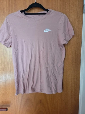 Rosa Nike t-shirt i bomull - Säljer en rosa t-shirt från Nike med klassisk logga på bröstet. T-shirten är i mjuk bomull och har en enkel, rund halsringning och korta ärmar. Perfekt för en avslappnad och sportig stil.