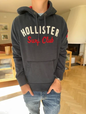 Hollister hoodie vintage herr mörkblå - Hollister Surf Club hoodie i mörkblå färg med snygg röd/vit brodyr framtill. Klassisk Hollister-design med huva och magficka. Stilren och lättmatchad modell i fint skick.  #Hollister #hoodie #surfclub #preppy #Y2K #streetwear #college #vintage