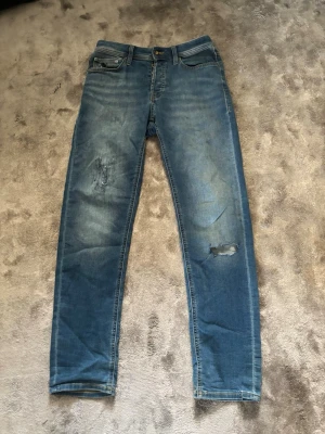 Blå slitna jeans - Säljer ett par blå skinny jeans med slitningar och hål på ena benet. Jeansen har klassisk femficksdesign, knappgylf och är tillverkade i ett stretchigt denimtyg för skön passform. Perfekta för en avslappnad och trendig look.