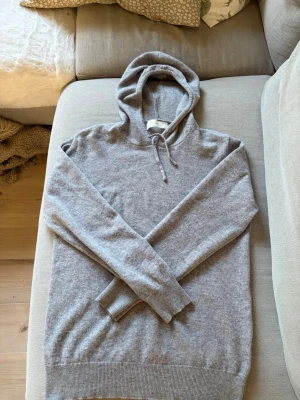 Grå cashmere hoodie från Sayless - Mysig grå hoodie i mjuk cashmere från Sayless. Klassisk modell med huva och dragsko, långa ärmar och ribbade muddar. Perfekt för en chill och stilren look. Passar dig som gillar både komfort och enkel stil.